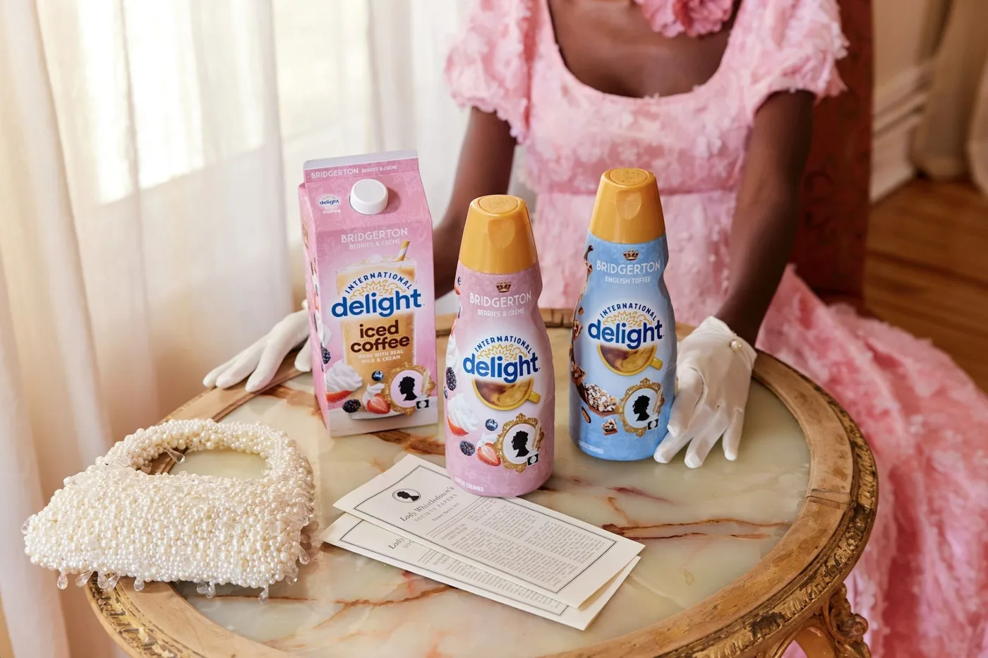 The limited-edition Bridgerton International Delight range - a pink Berries & Crème carton and bottle and a blue English Toffee bottle - staged on a marble table beside a pearl handbag and a Lady Whistledown society paper.